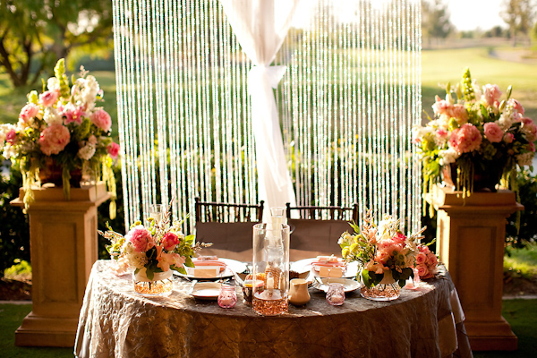 Beautiful sweetheart's table decor at outdoor reception - dark gold tablecloth with ivory candle in the center and dark pink, orange, coral, yellow, and light green floral arrangements on each side of the table and two larger arrangements on pedestals on each side of the table with crystal beads hanging in the background - wedding photo by Michael Norwood Photography